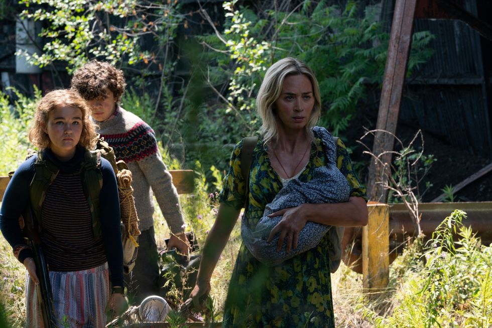 Noah Jupe, Millicent Simmonds and Emily Blunt return as the Abbott family in "A Quiet Place Part II." It's a tight angle shot of the actors walking through a sunny field surrounded by overgrown brush and grass. Blunt carries a baby in her left arm.