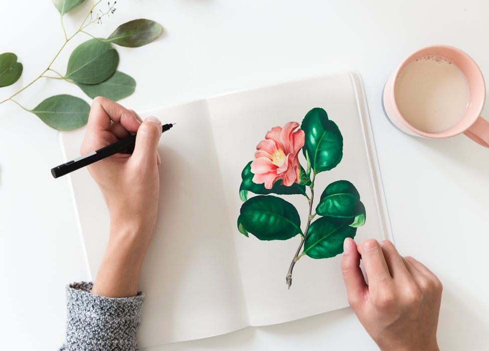 Person drawing a pink flower in a sketchbook next to a cup of milk and green leaves.