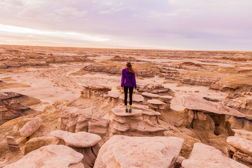 Person in purple jacket standing on rocky formations in a vast desert landscape at twilight.