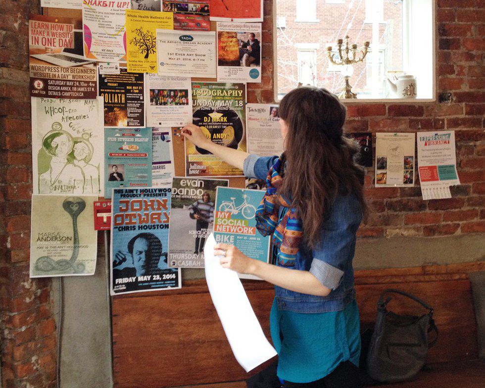Person pinning posters to a busy bulletin board on a brick wall.