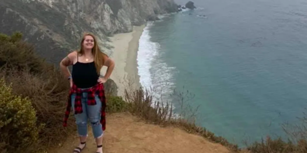 person smiling at camera with mountains and blue water behind her