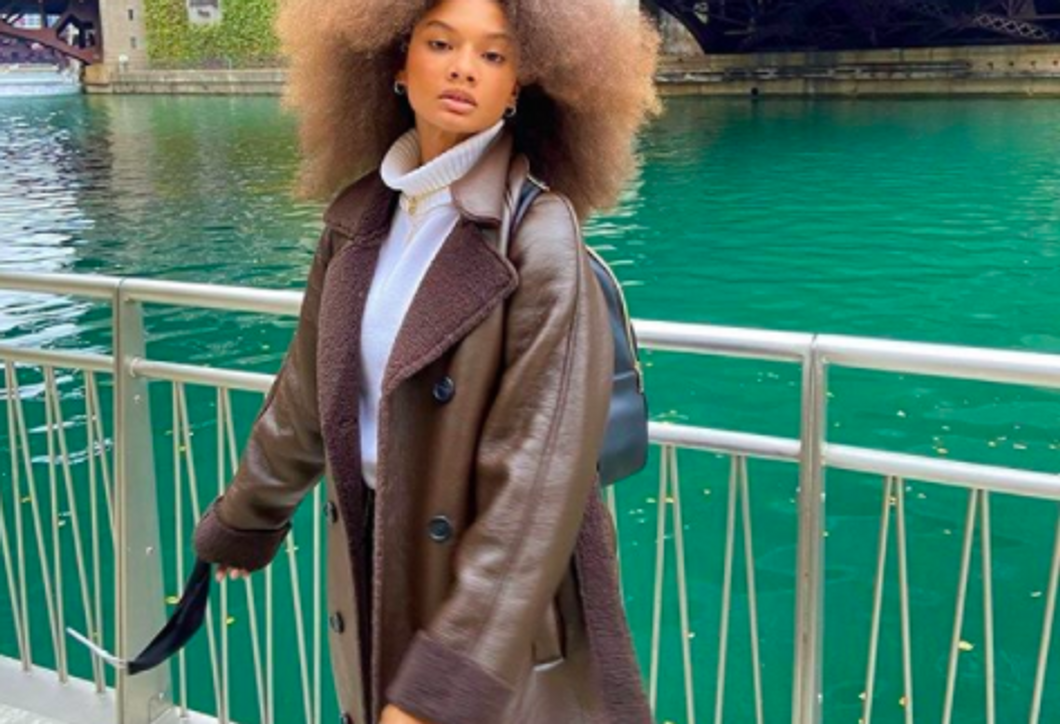 person wearing faux fur-lined brown leather coat and white turtleneck standing in front of green river