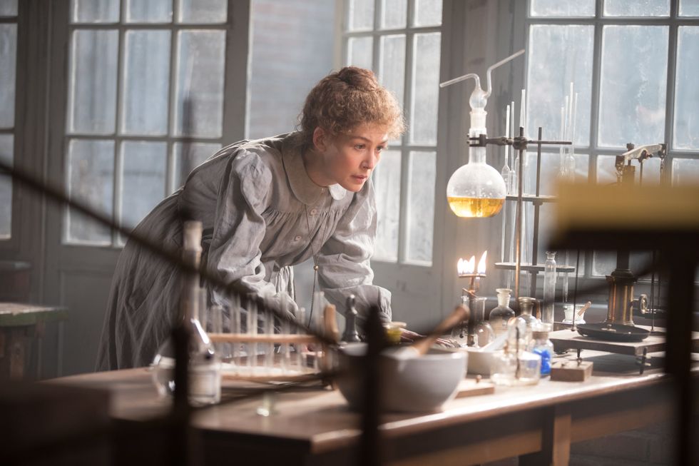 Rosamund Pike is physicist and chemist Marie Curie. She is wearing a long-sleeved grey dress and staring at a burner as it boils a beaker from a chemistry set.