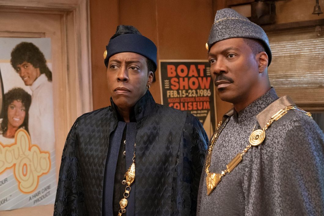 Semmi (left, played by Arsenio Hall) and Prince Akeem (Eddie Murphy) stand side by side in a barbershop in Queens, New York. Both men are wearing regal coats, clothes and giant gold royal necklaces. Arsenio is wearing a black chevron coat over navy clothing while Eddie wears a grey coat. Both wear hats that match their respective outfits.