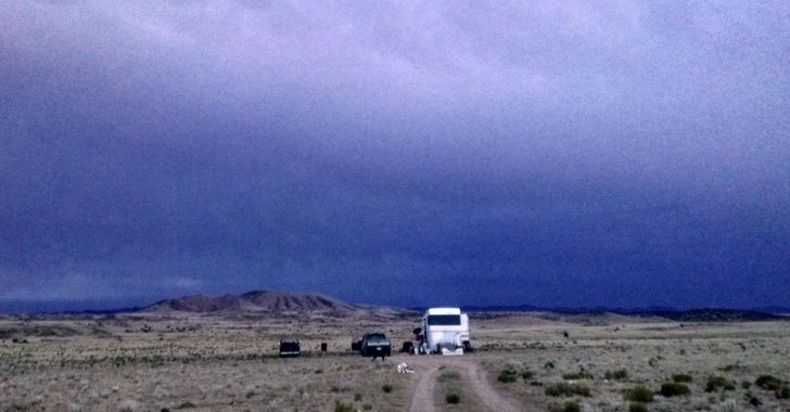 Several vehicles and a fifth wheel at the end of a dirt road on a barren plain with a dark cloudy sky and hill in the background