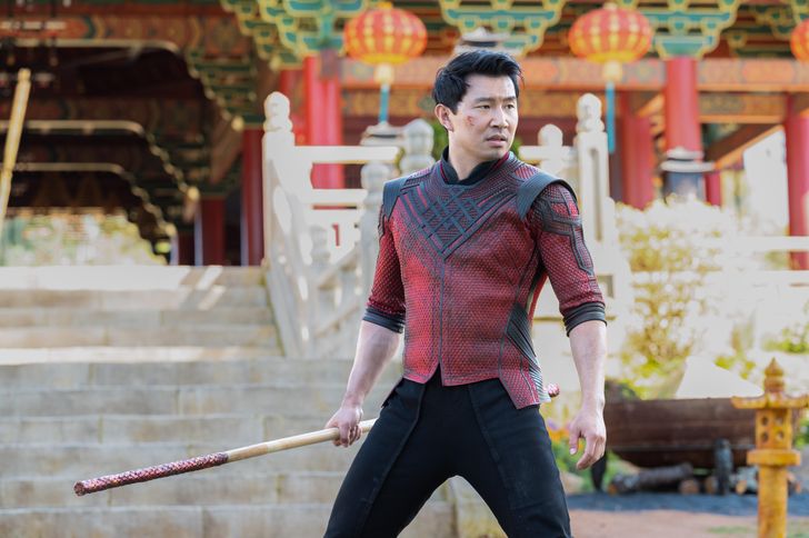 Simu Liu (as Shang-Chi) stands in front of a temple with red pillars. He's holding a bowstaff, and he's wearing red armor made from dragon scales.