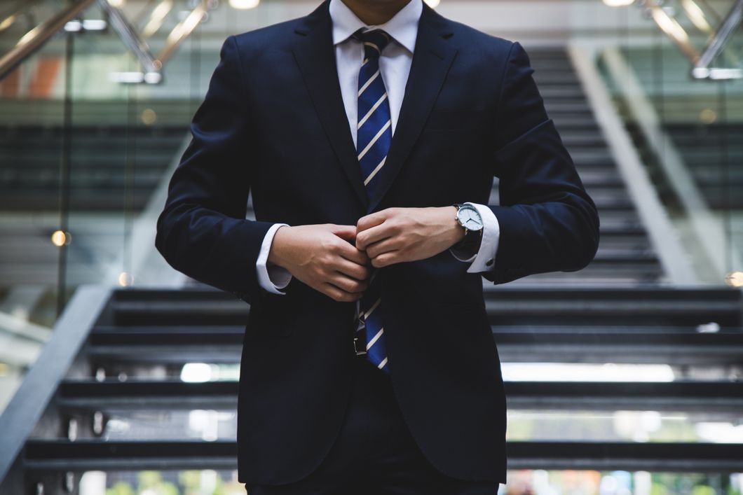 Someone buttoning their suit jacket, wearing a blue tie in front of stairs.