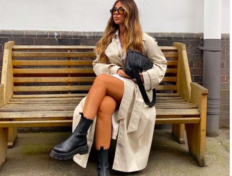 someone sitting on bench wearing black sunglasses, a tan trench coat, and black Chelsea boots while holding a black purse