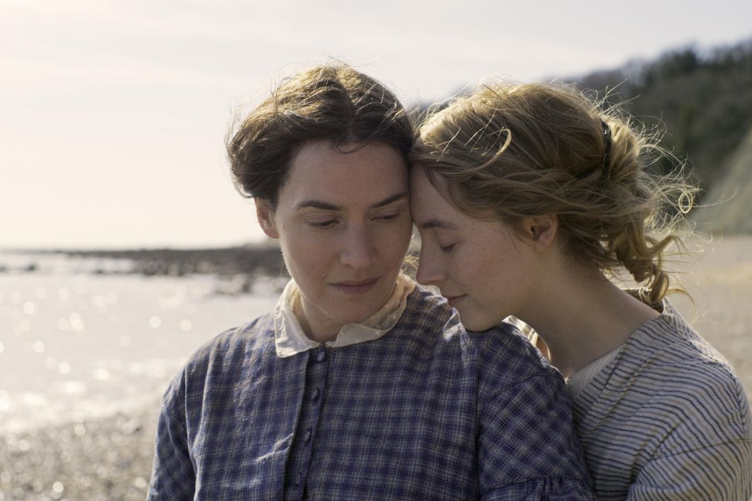 The photo is a close up shot of Kate Winslet (left) as Mary Anning and Saoirse Ronan (right) as Charlotte Murchison. Saoirse has her forehead to the side of Kate's and Saoirse rests her chin on Kate's left shoulder, with her eyes closed. Both are wearing casual period clothing from the late 1800s England. The Southern Shore is shown, blurred, in the background.