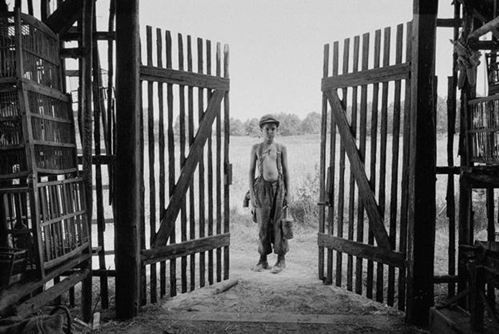 The photo is in black and white. A young boy in the middle of Eastern Europe at the end of World War Two stands between two tall wooden gates. He's shirtless and wearing suspenders with a newsboy cap.
