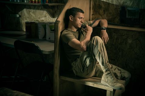 Tom Holland as "Cherry" sits near a phone booth as he calls Emily (Ciara Bravo) while stationed in Iraq with the United States Army. He wears a tan shirt, army camoflauge pants and high-lace boots.