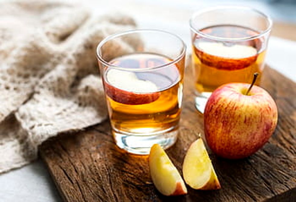 Two clear glasses of apple cider with a piece of an apple in each one, on top of a wood board with a full apple next to them