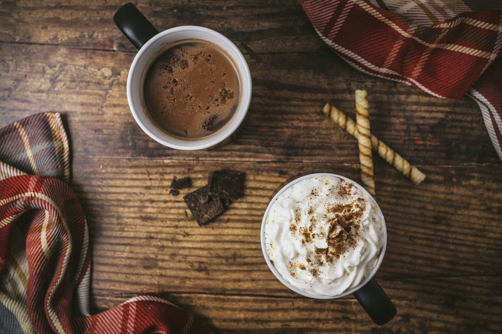 Two mugs of hot chocolate, one with whipped cream, on a wooden surface decorated with chocolate and a flannel scarf.