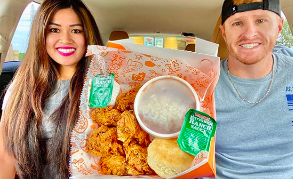 two people sitting in car, smiling and holding up a basket of chicken wings with a biscuit and ranch sauce on the side