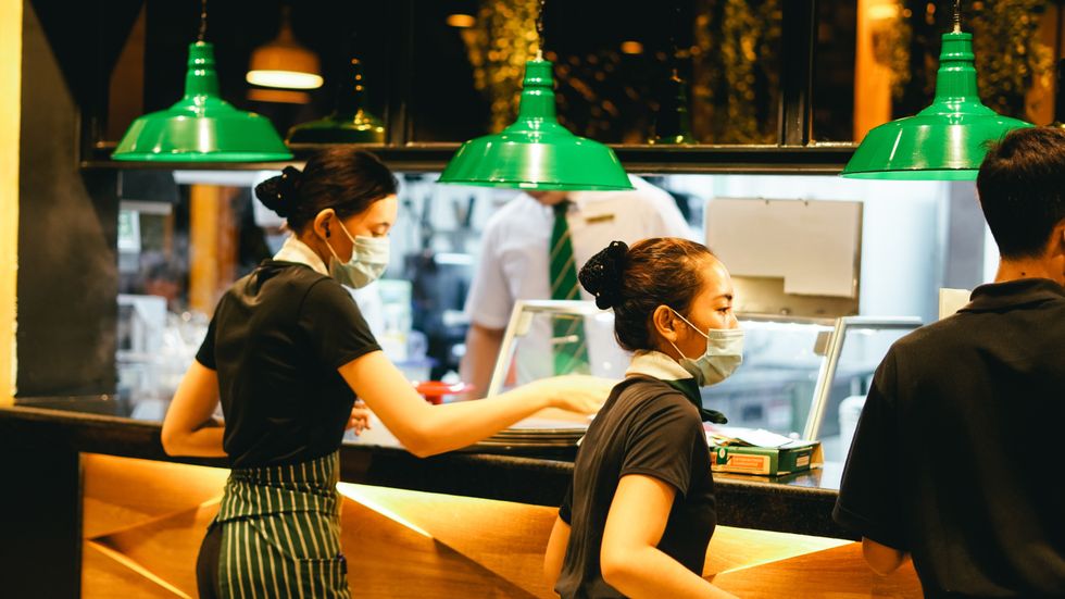 two women wearing aprons, all black, and masks with hair in buns standing behind plexi glass