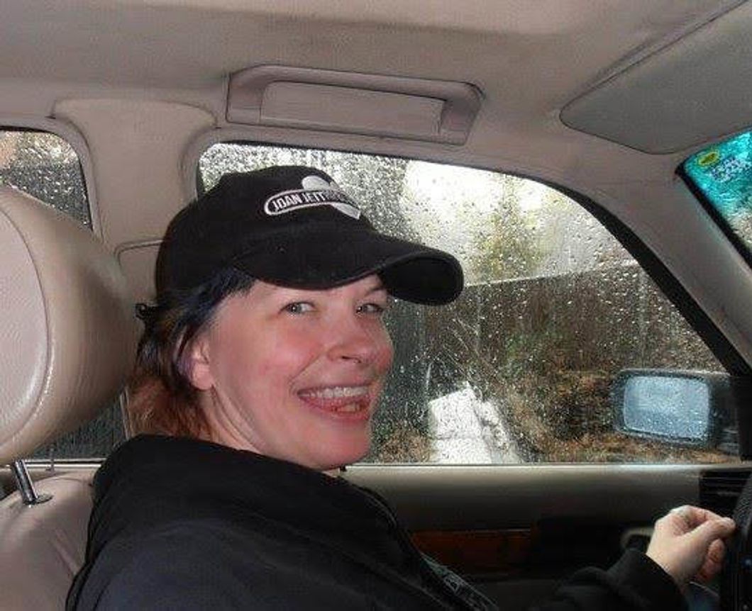 Woman in a black Joan Jett baseball hat biting her tongue between her teeth sitting in a car with rain outside the window