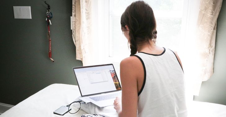 Woman in white tank top sitting on bed in front of laptop computer