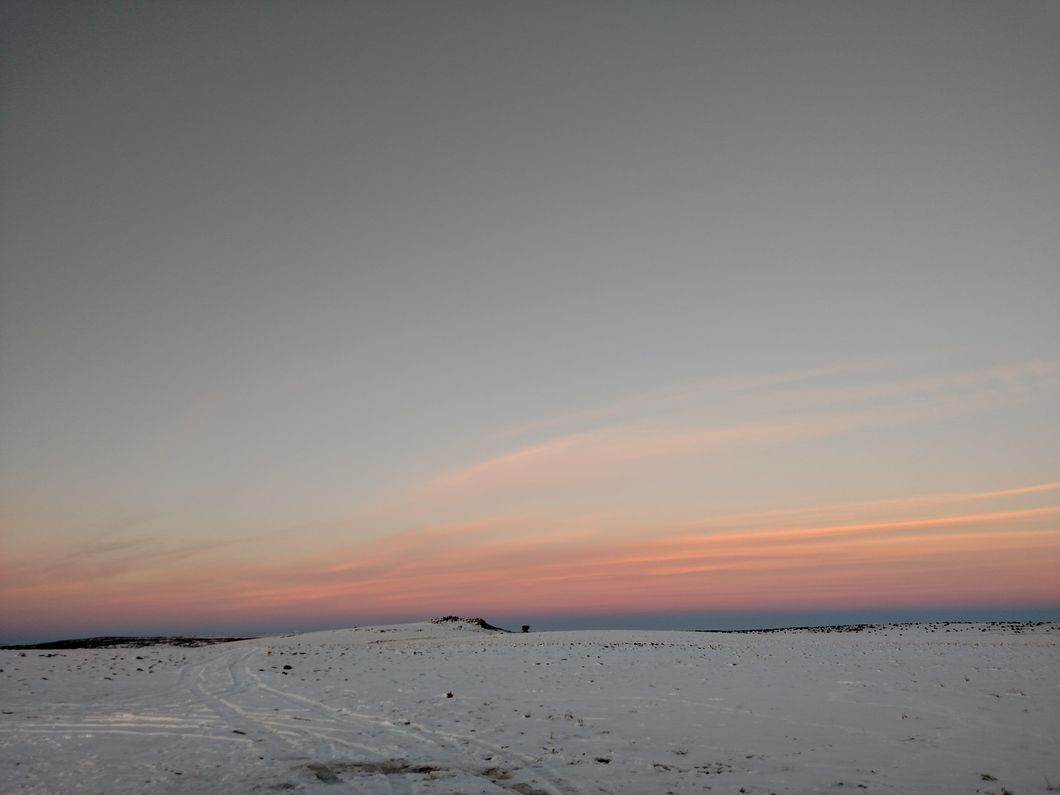 Yellow pink and purple sunset over a barren landscape covered in snow with tire tracks leading away from the source